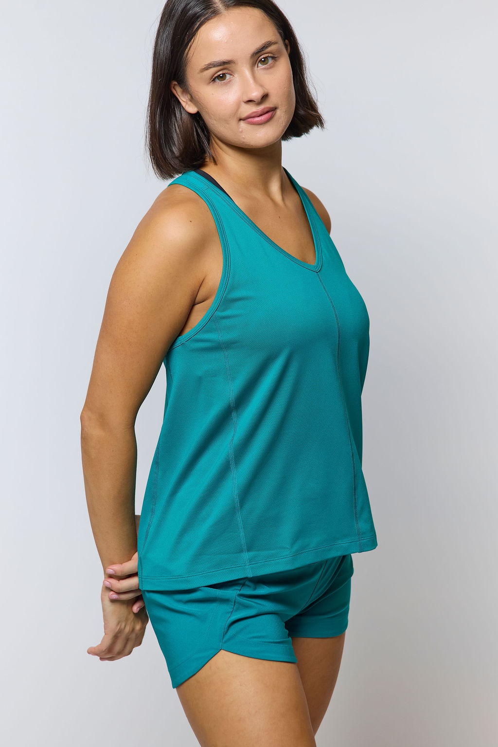 Woman wearing teal workout tank top and matching shorts, hands clasped behind her back.