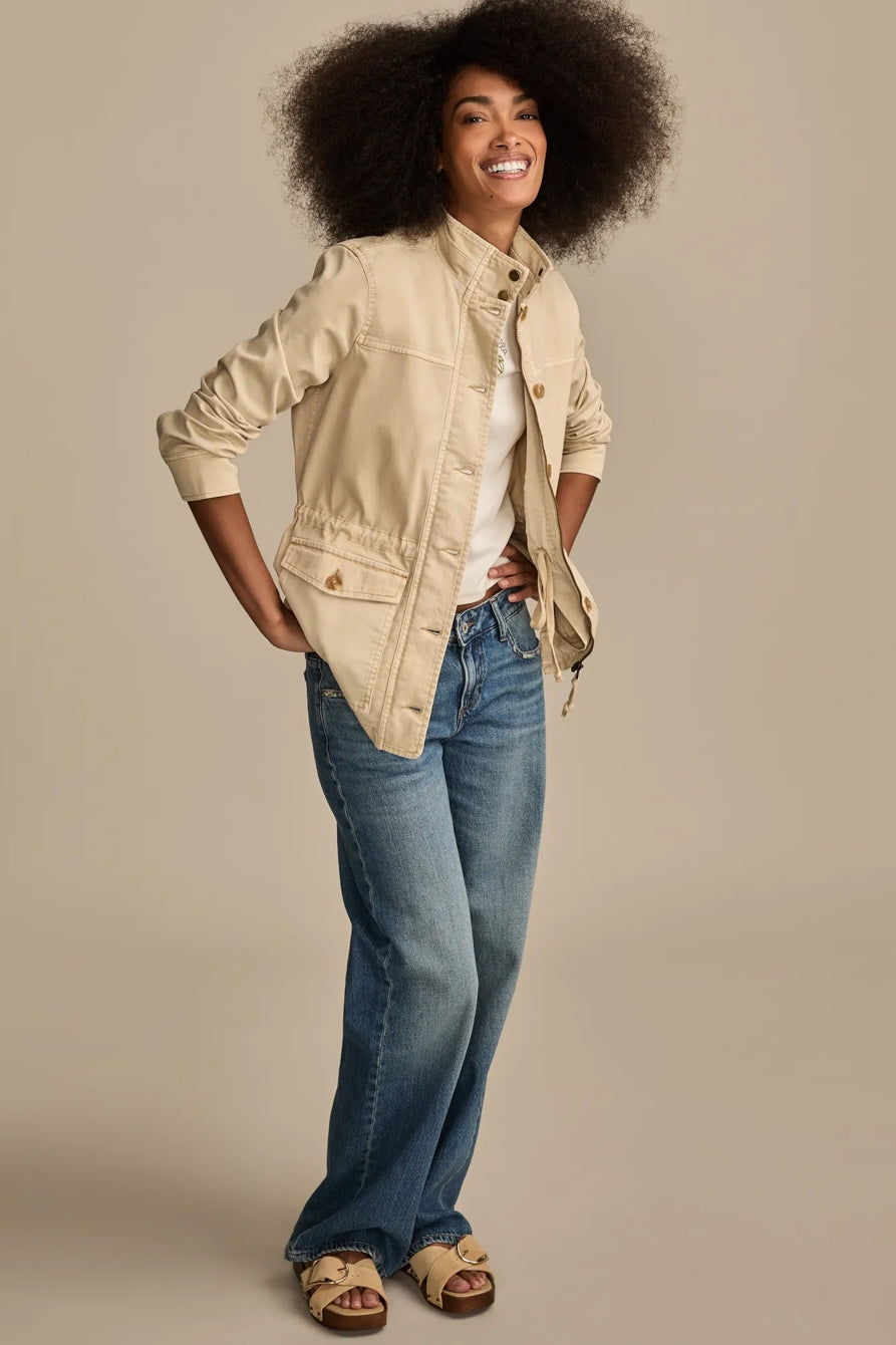 Woman wearing a tan utility jacket and blue jeans, hands on hips.