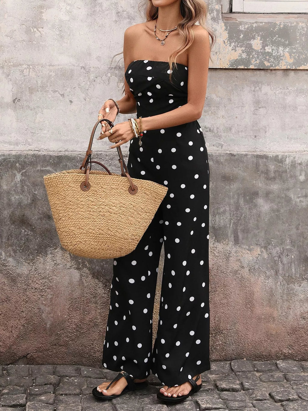 Woman wearing a black strapless wide-leg jumpsuit with white polka dots, holding a straw tote bag.