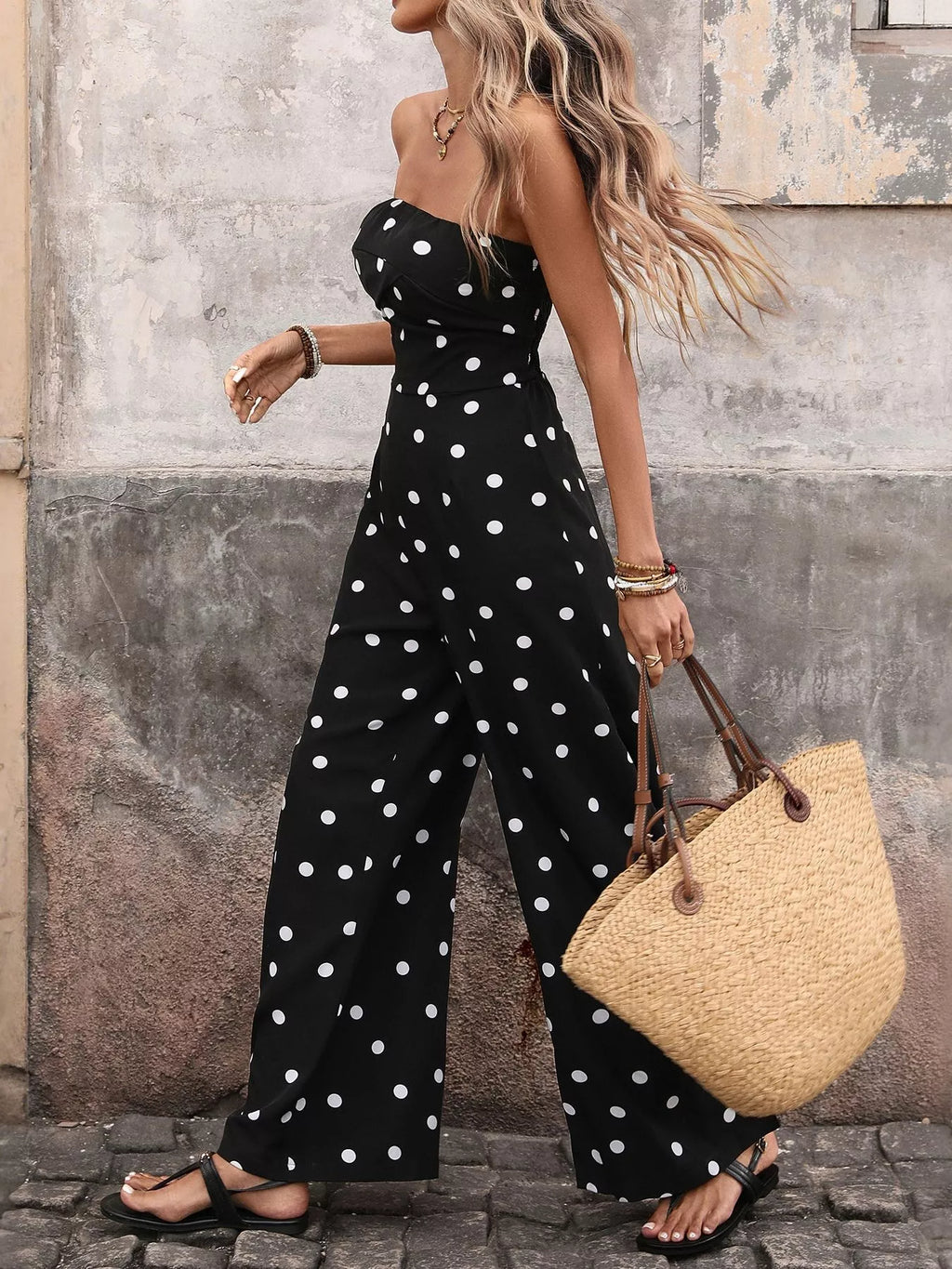 Woman wearing a black strapless wide-leg jumpsuit with white polka dots and carrying a straw bag.