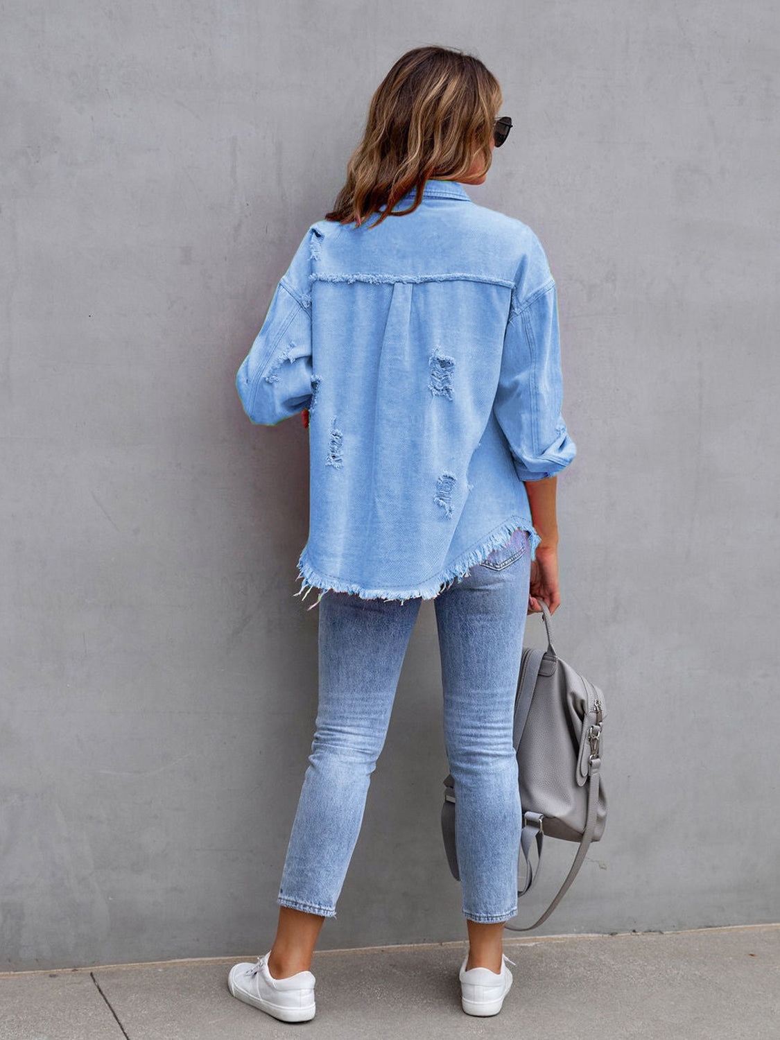Back view of a light blue distressed drop shoulder denim jacket with fringe hem and ripped details.