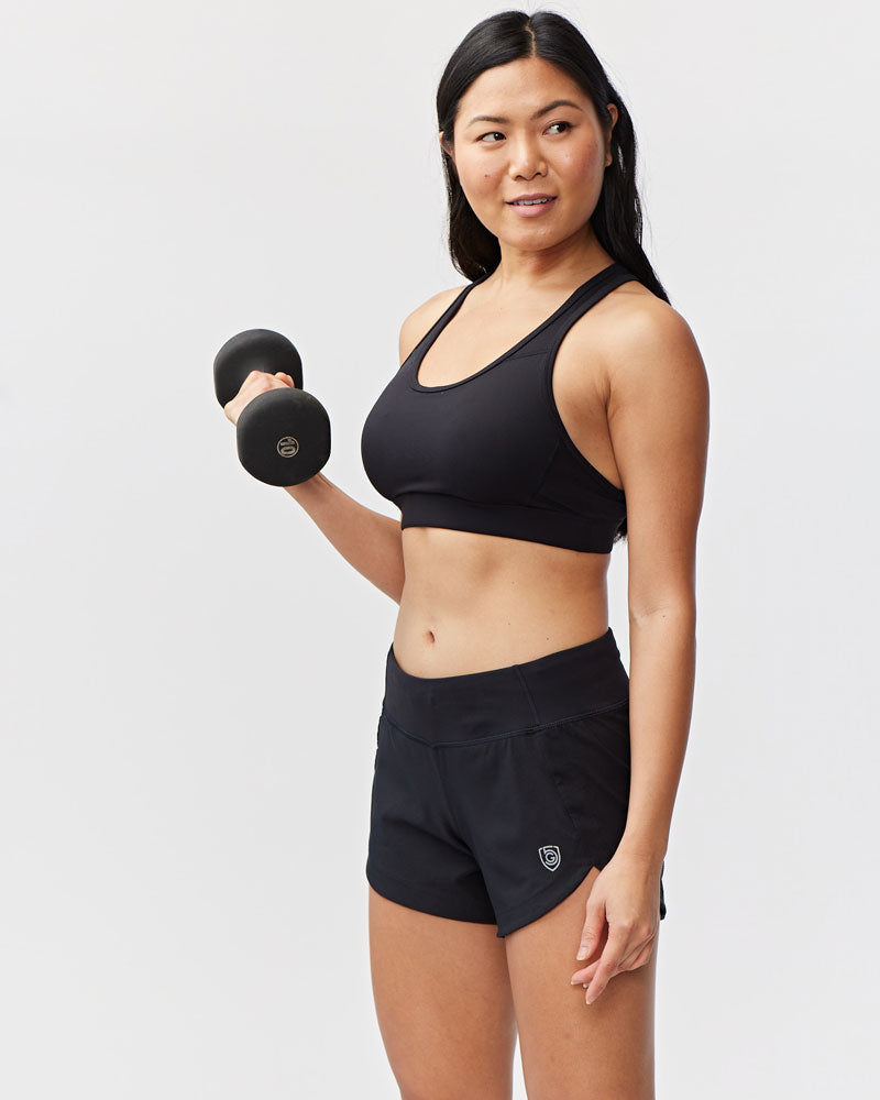 Woman in black Goal Five Excel workout shorts and sports bra holding a dumbbell.