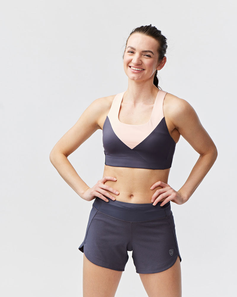Woman wearing charcoal grey workout shorts with a pink and grey sports bra, hands on hips.