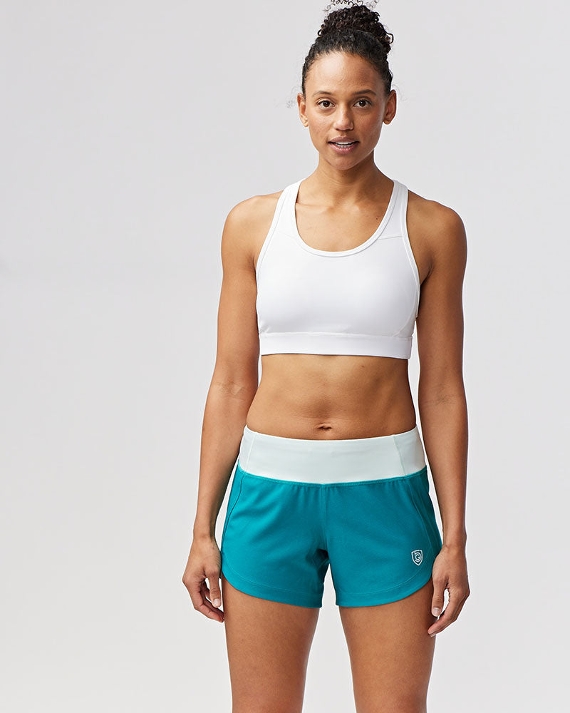 Woman wearing white sports bra and teal Goal Five Excel workout shorts with a light blue waistband.