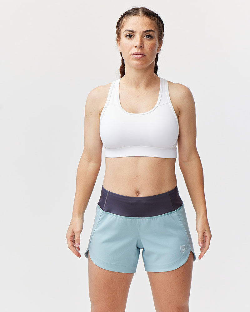 Woman wearing white sports bra and light blue Goal Five Excel workout shorts with a dark gray waistband.