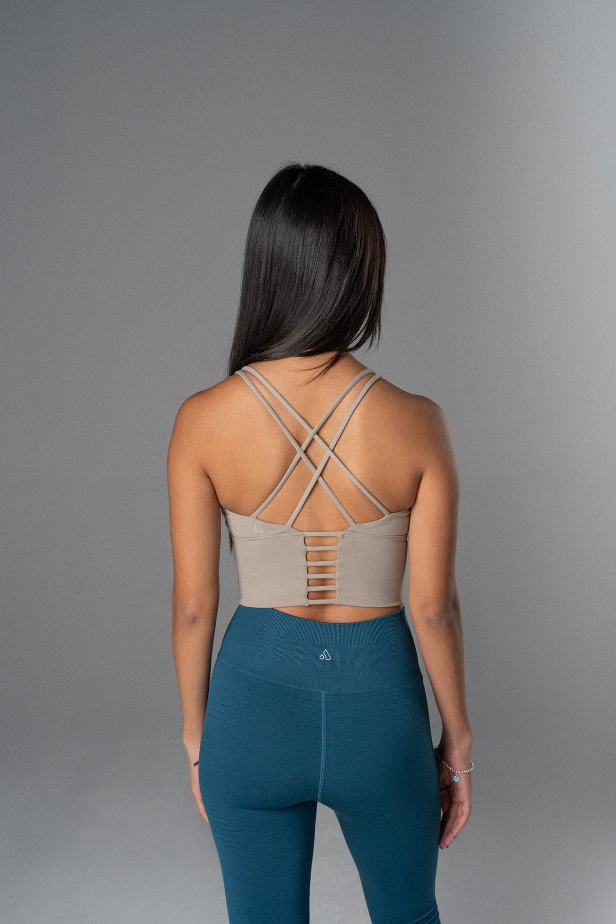 Back view of a woman wearing a beige Alyth Active Rebellious Crop Bra with criss-cross straps and blue leggings.