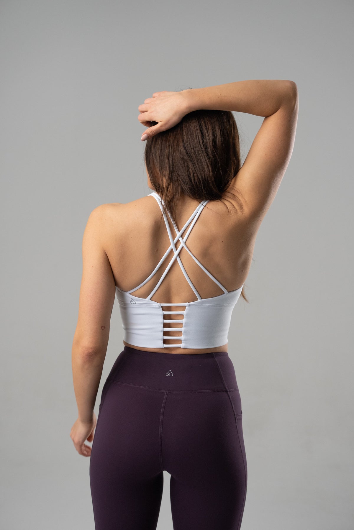 Woman from behind wearing a white high-impact crop bra with strappy back and purple leggings.
