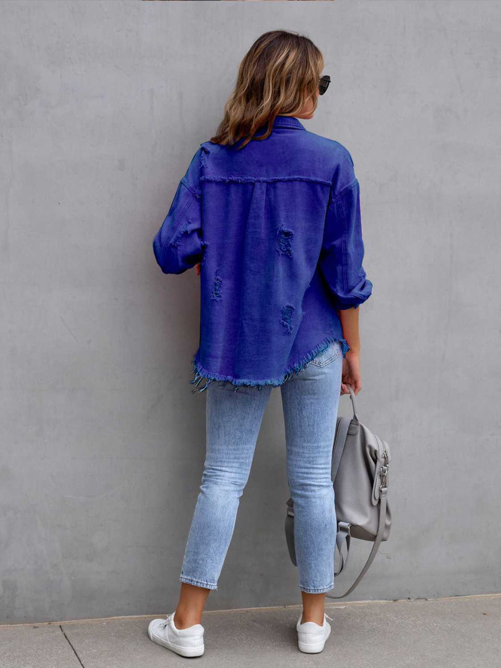 Back view of a woman wearing a royal blue distressed drop shoulder denim jacket with fringe hem.