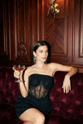 Woman in a black strapless lace corset dress holding a champagne glass, seated on a velvet couch.