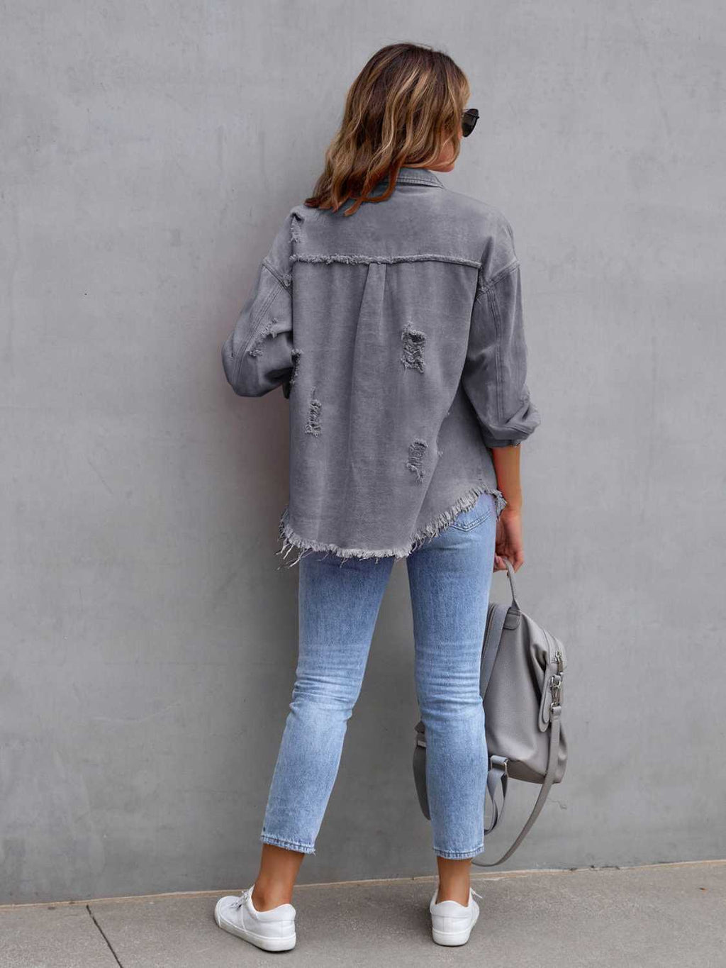 Back view of a woman wearing a grey distressed drop shoulder denim jacket with fringe hem.