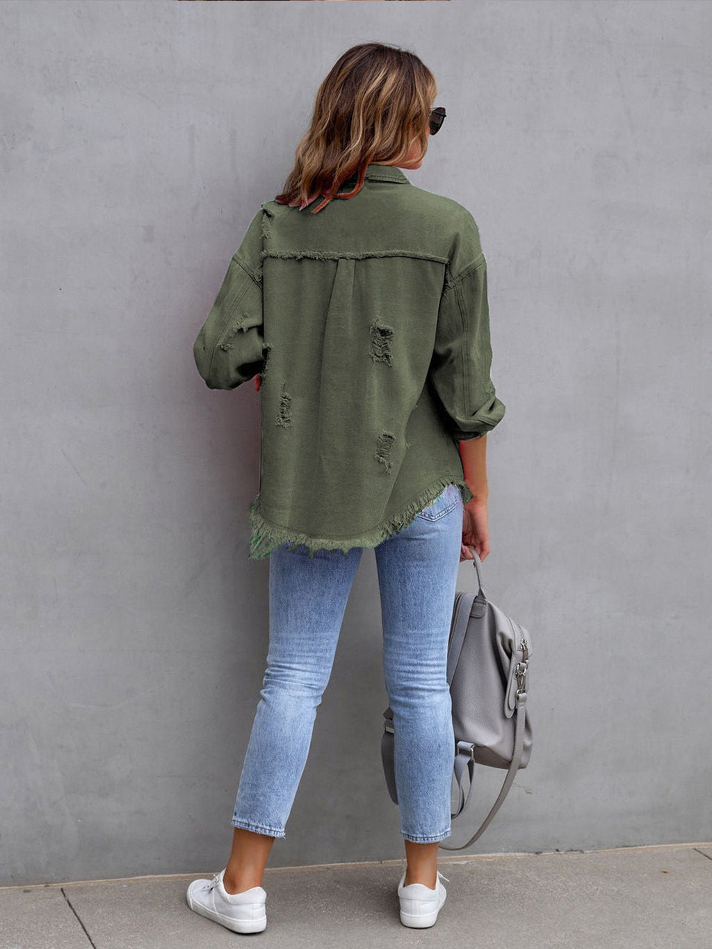 Back view of a woman wearing an olive green distressed drop shoulder denim jacket with fringe hem.