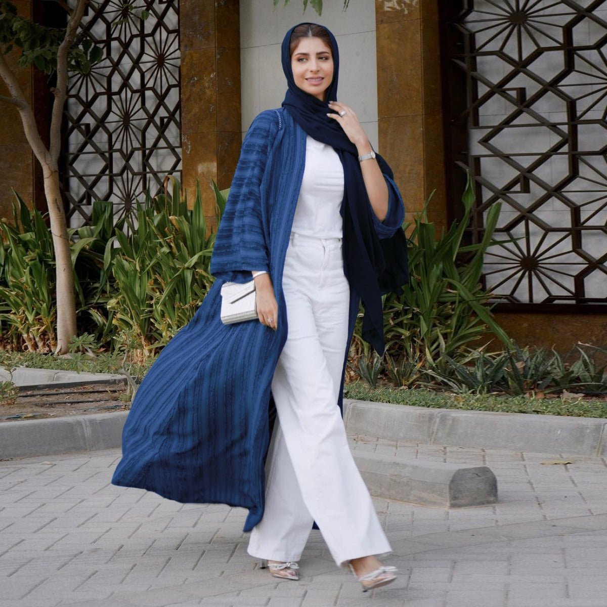 Woman wearing a blue vintage knitted longline open front cardigan abaya over white pants and top.