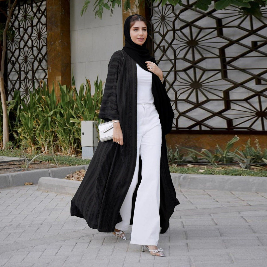 Woman wearing a black vintage knitted longline open front cardigan abaya over white pants and top.