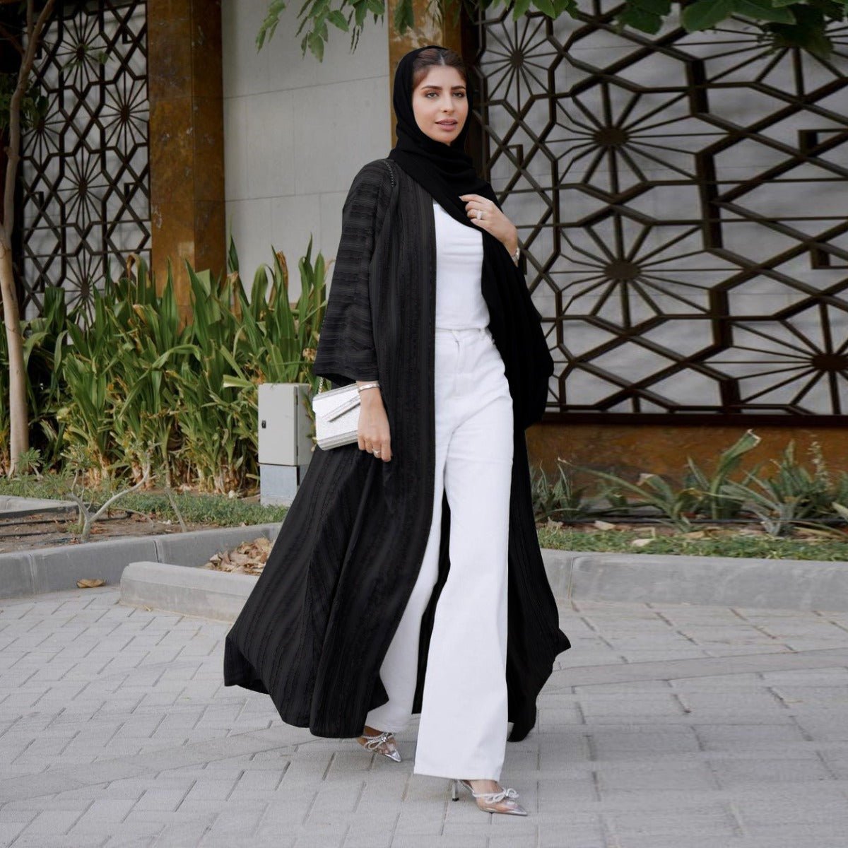 Woman wearing a black vintage knitted longline open front cardigan abaya over white pants and top.