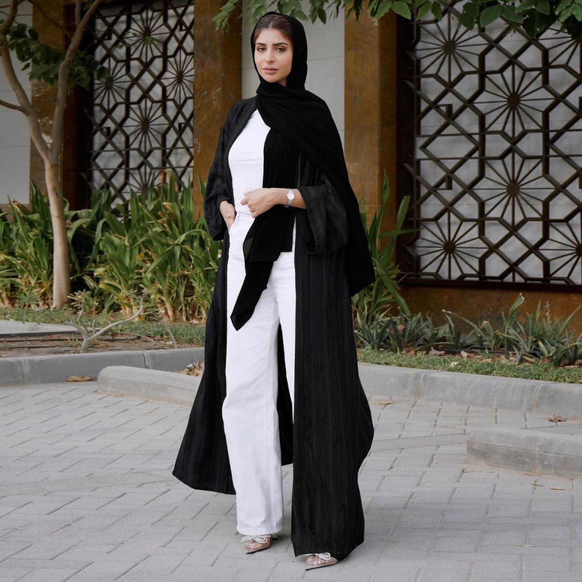 Woman wearing a black vintage knitted longline open front abaya over a white top and pants.